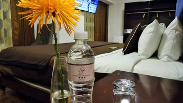 A hotel room with a bed, a TV, a bottle of water, a vase with a flower, and a glass of water on a table.