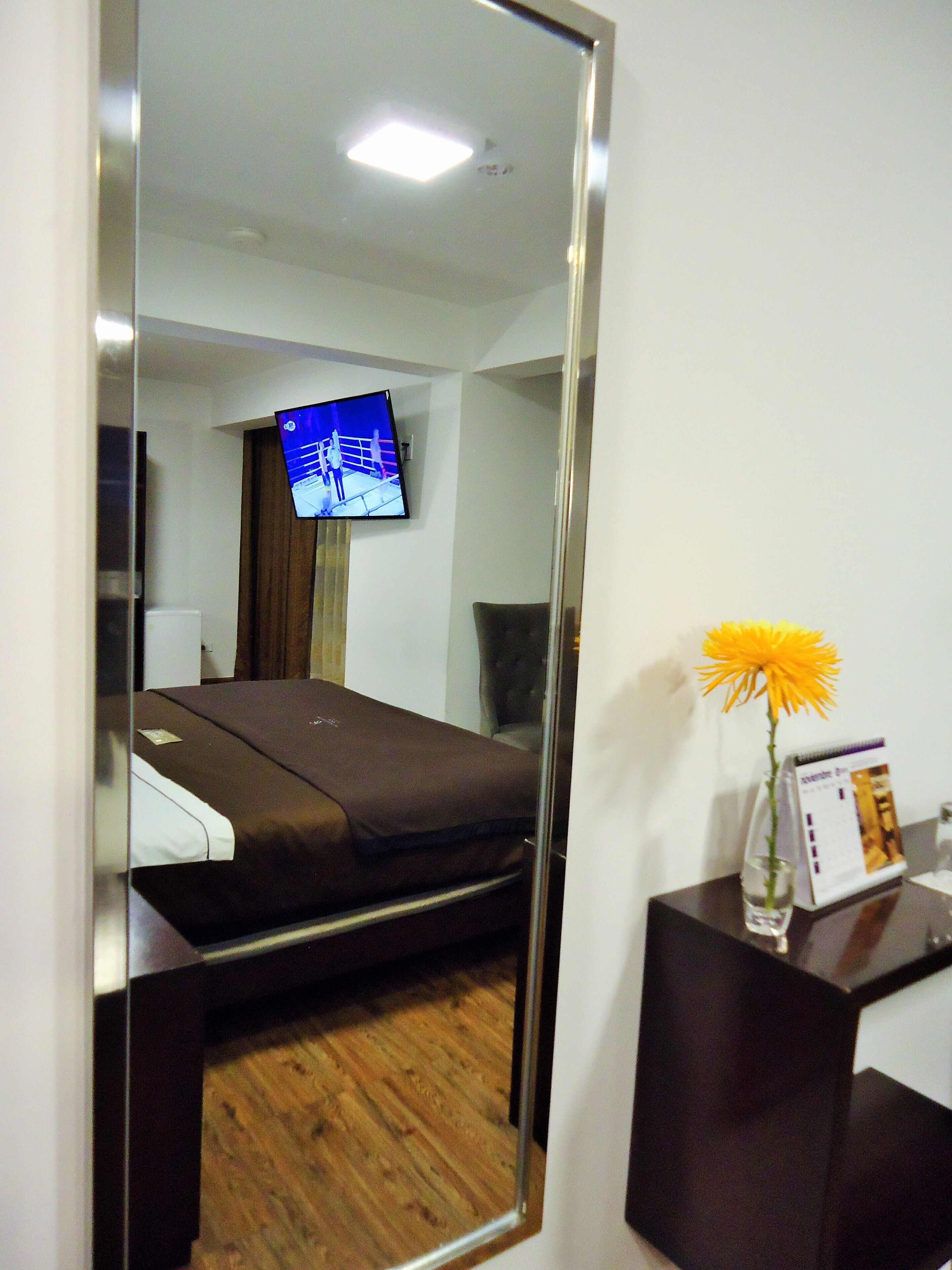 A hotel room with a bed, a television mounted on the wall, a small table with a vase of flowers, and a brochure on the table.