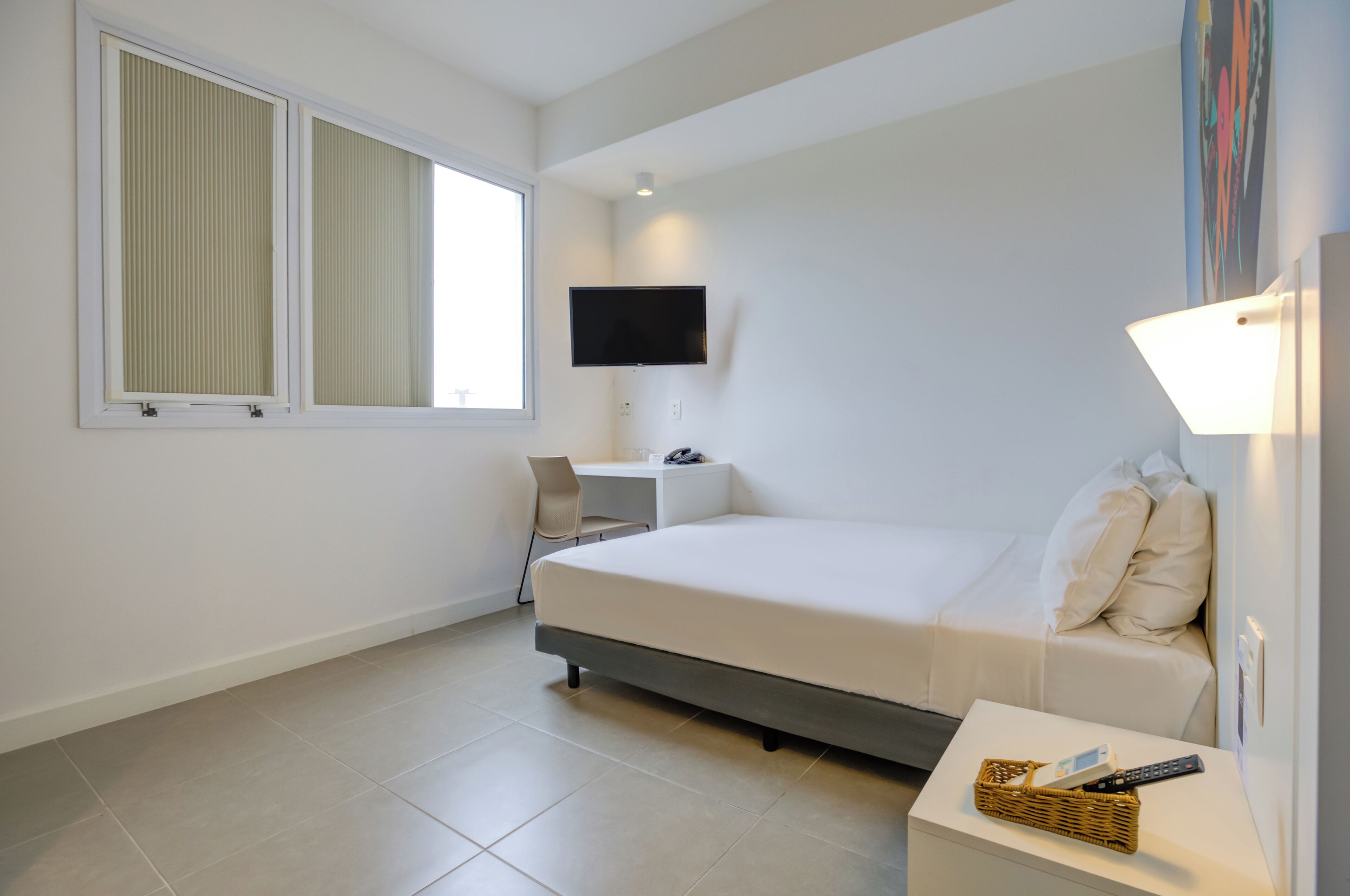 Quarto de hotel moderno com cama, escrivaninha, cadeira, televisão e janela.
