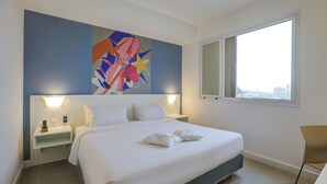 Quarto de hotel moderno com uma cama grande, duas mesas de cabeceira, um criado-mudo, uma cadeira e uma janela ampla com vista para a cidade.