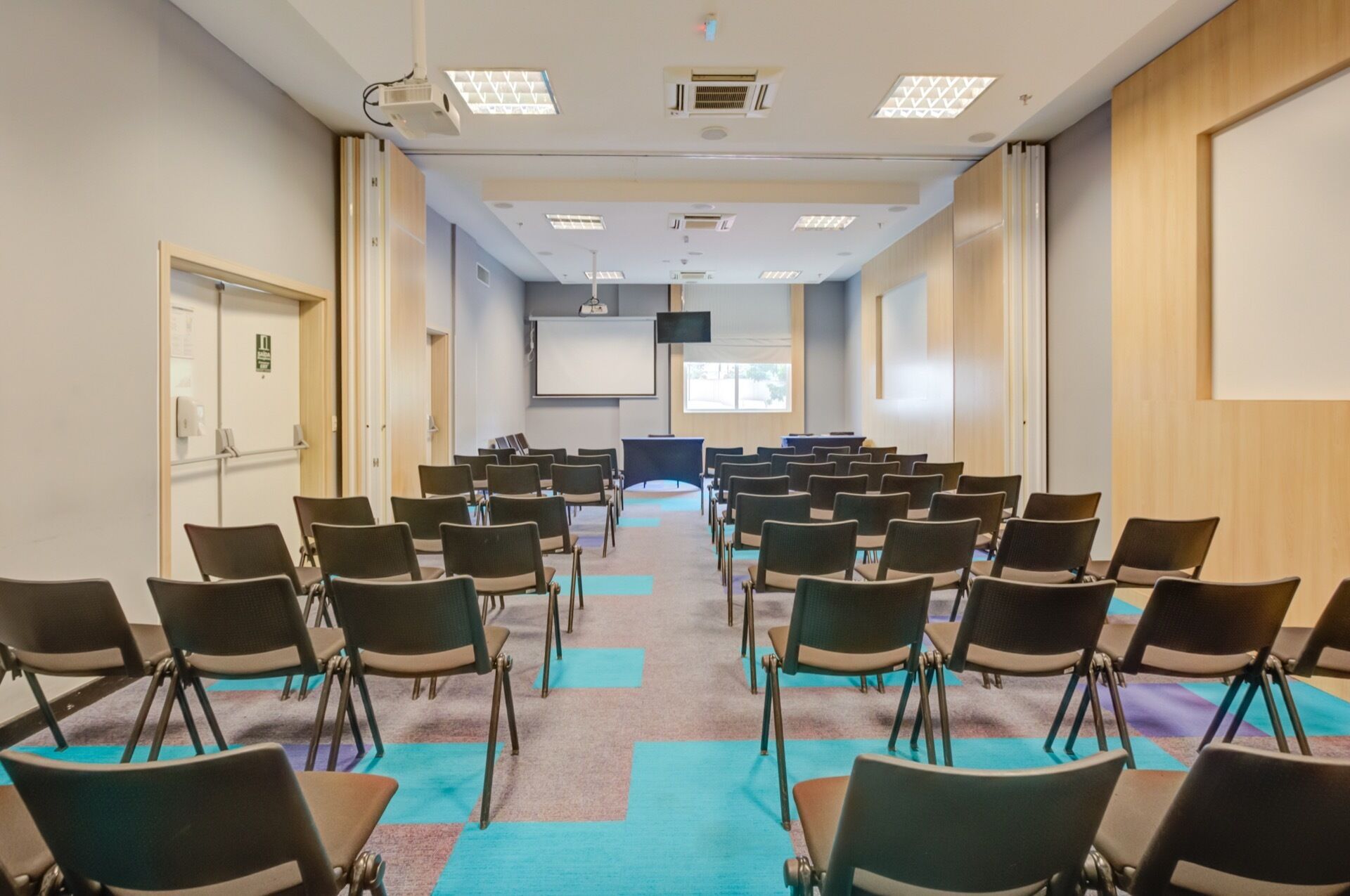 Sala de conferências com fileiras de cadeiras voltadas para uma tela de projeção, equipada com projetor e ar condicionado.