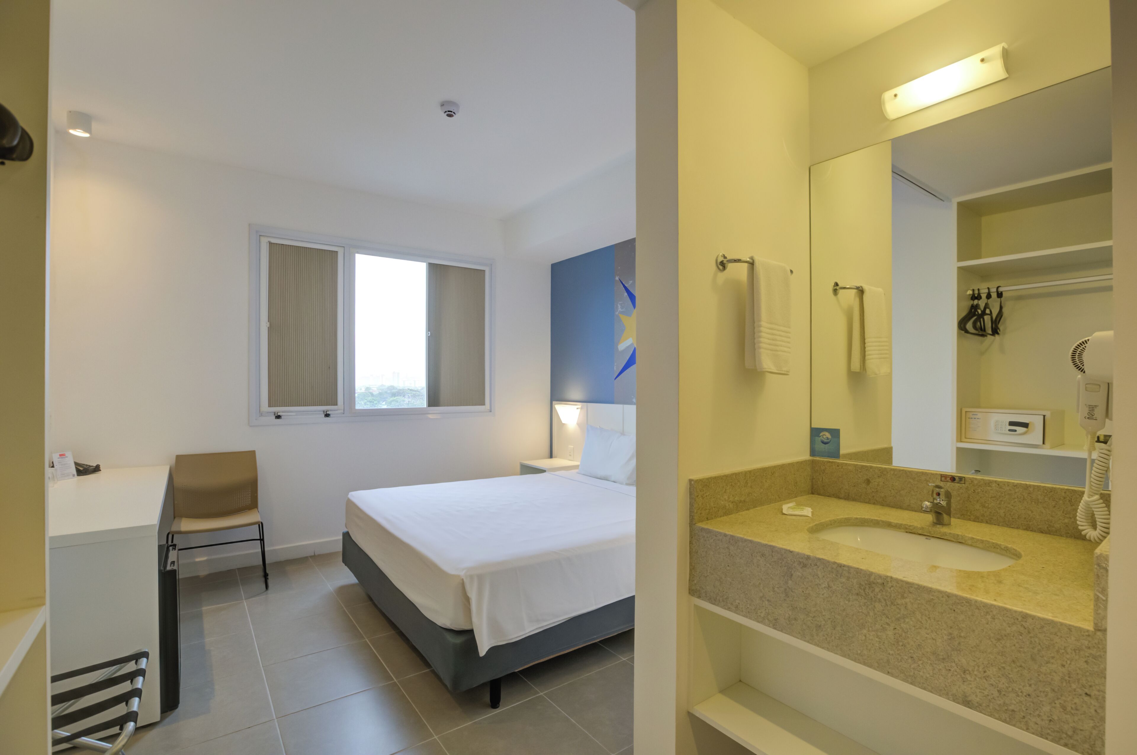 A hotel room with a bed, a desk, a chair, a bathroom with a sink and mirror, and a window.