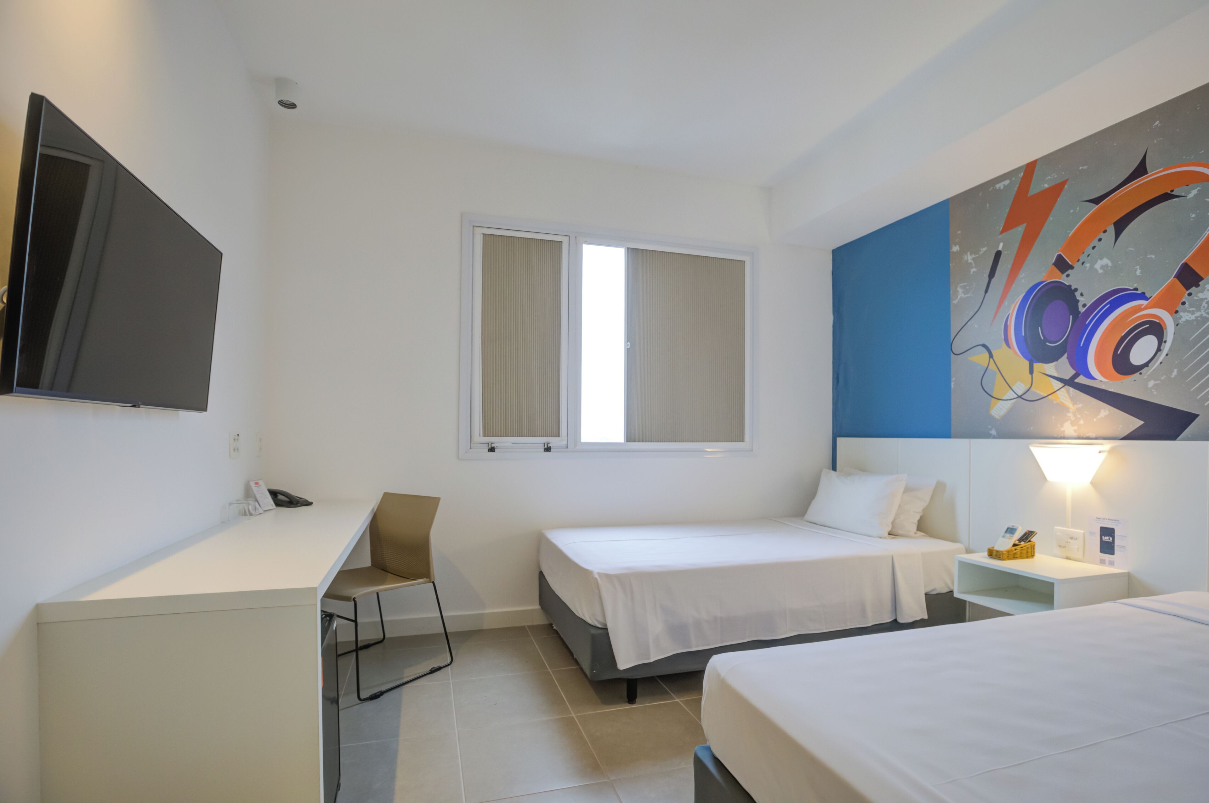 Quarto de hotel com escrivaninha, duas camas, uma cadeira, televisão e um quadro na parede.