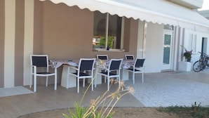 Un espacio al aire libre cubierto con una mesa y sillas, una bicicleta estacionada afuera, y un edificio con ventana y persianas.