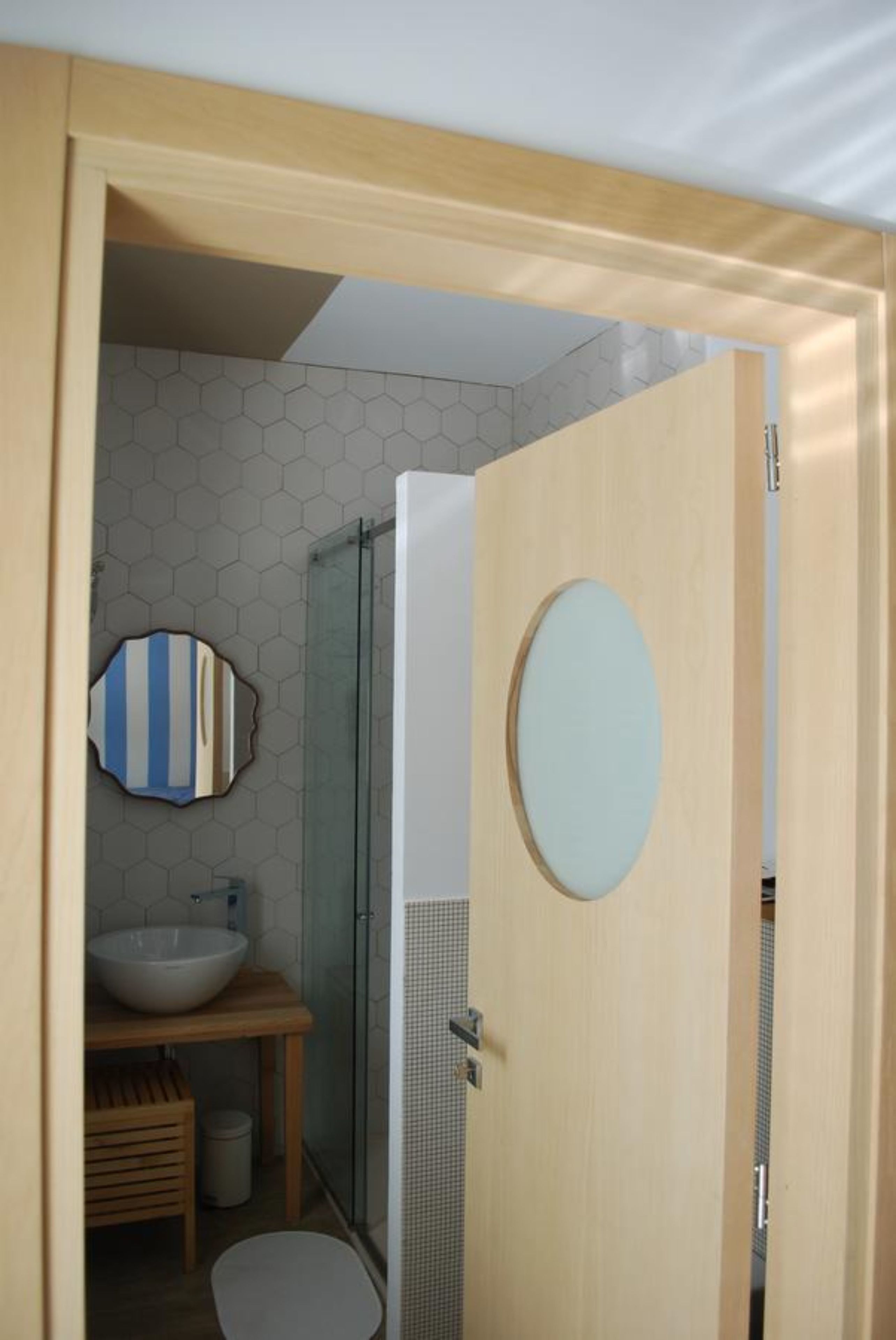 Un baño moderno con puerta de madera, espejo redondo, cabina de ducha de vidrio y un salpicadero de azulejos hexagonales.