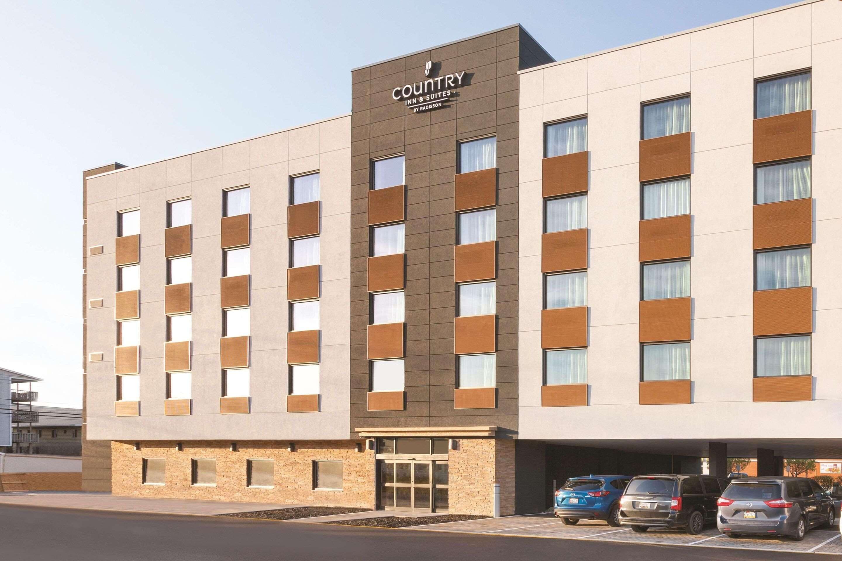 A modern hotel building with a flat facade, multiple windows, and a parking area with several cars.