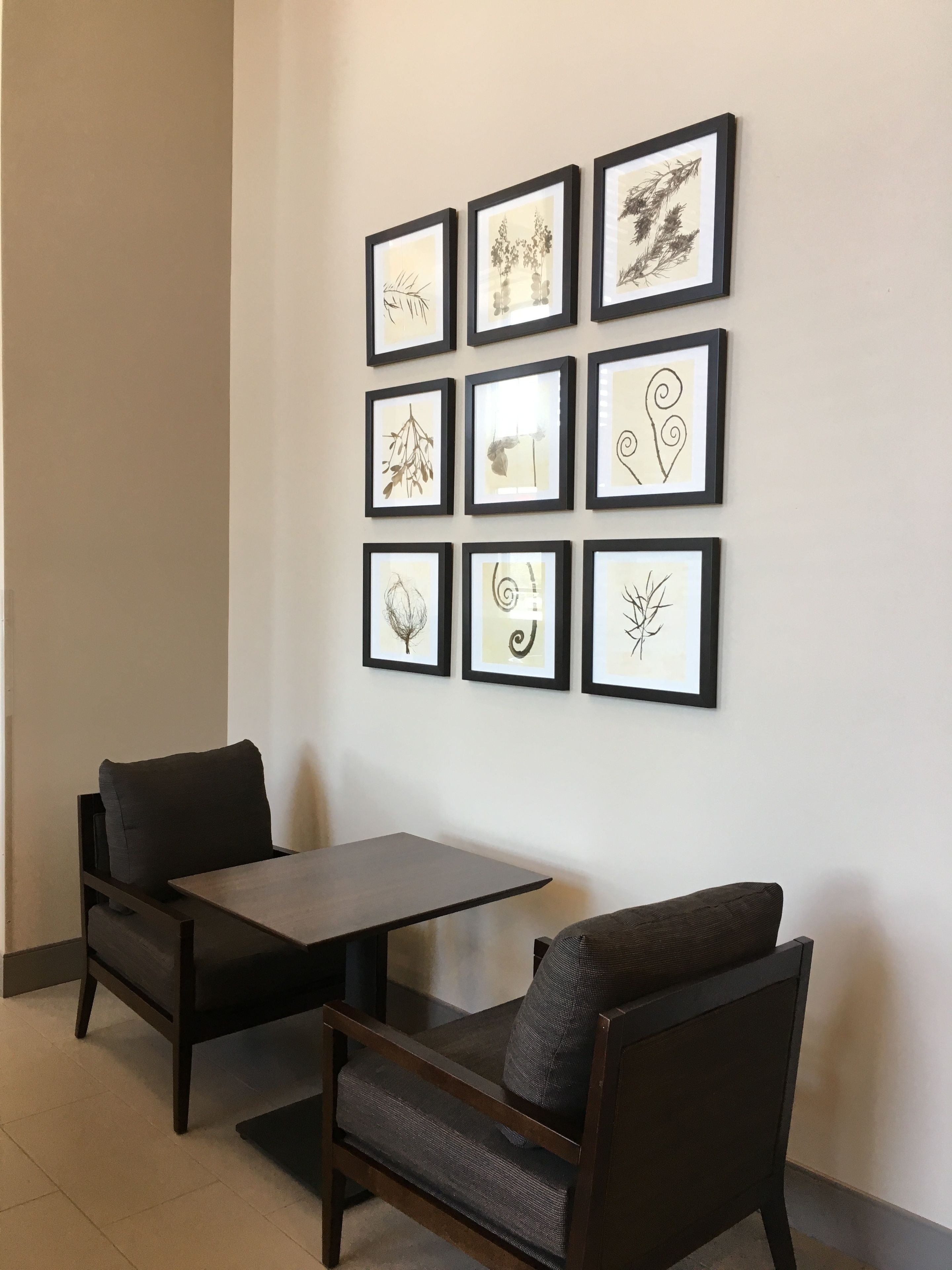 A room with a dark wooden table and two chairs, a white wall with nine framed botanical prints, and a small window.