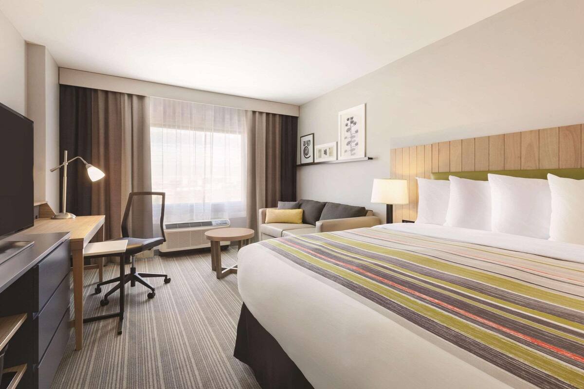 A hotel room with a large bed, a desk, a chair, a small round table, a TV, and a window with curtains.