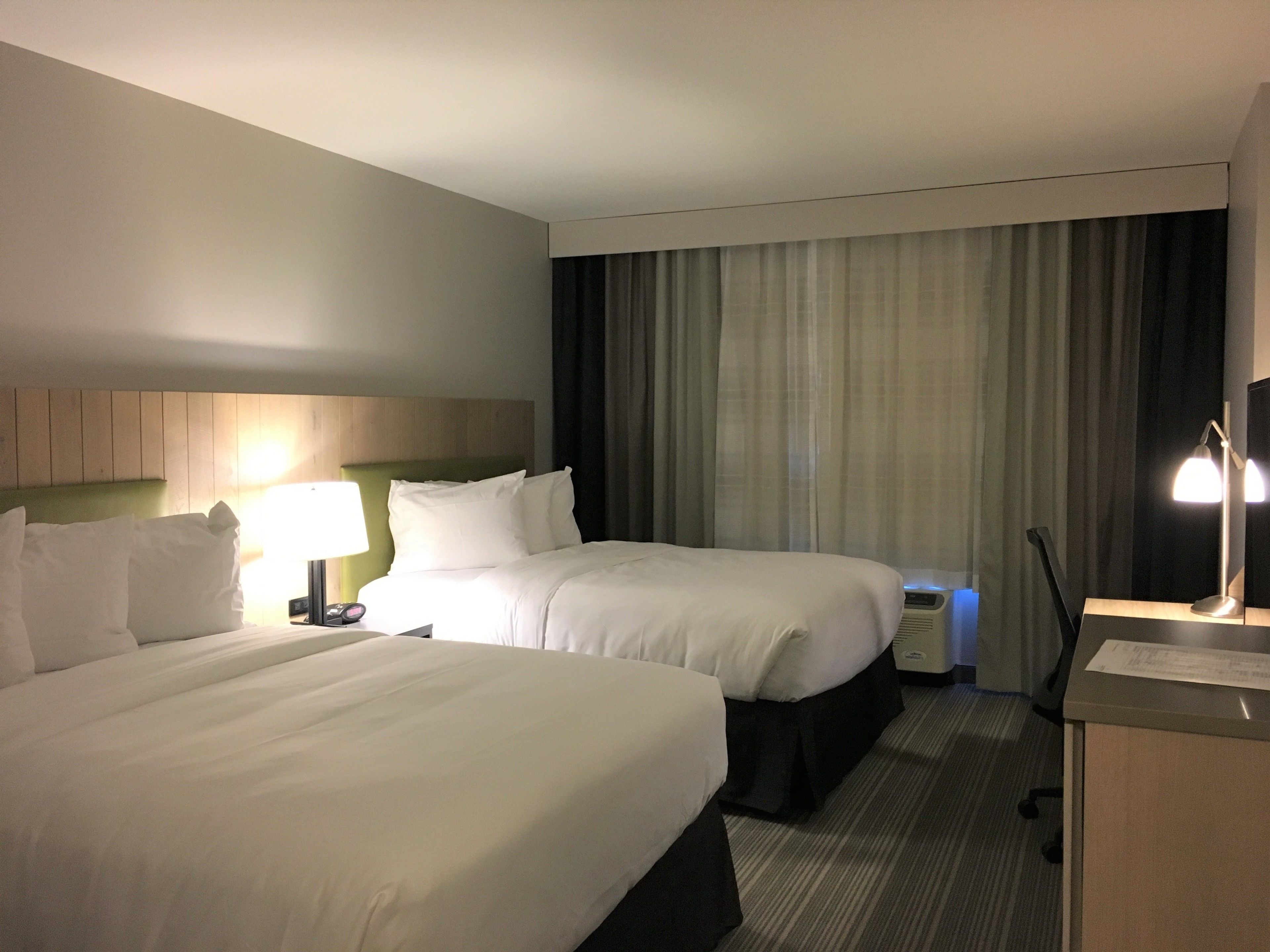 A hotel room with two beds, a desk, a lamp, and a window with curtains.