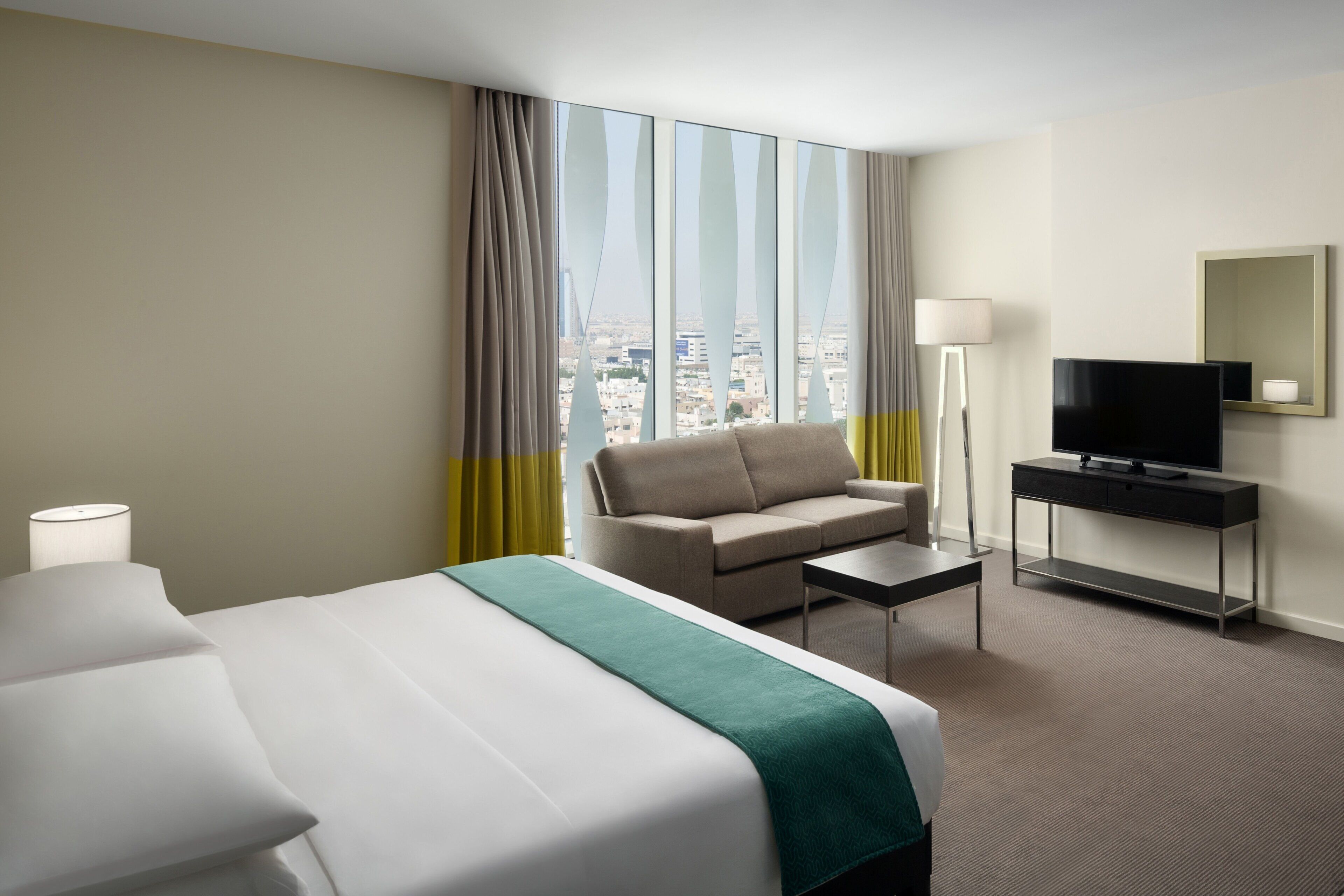 A hotel room with a bed, a sofa, a TV on a stand, and a view of the city through large windows.
