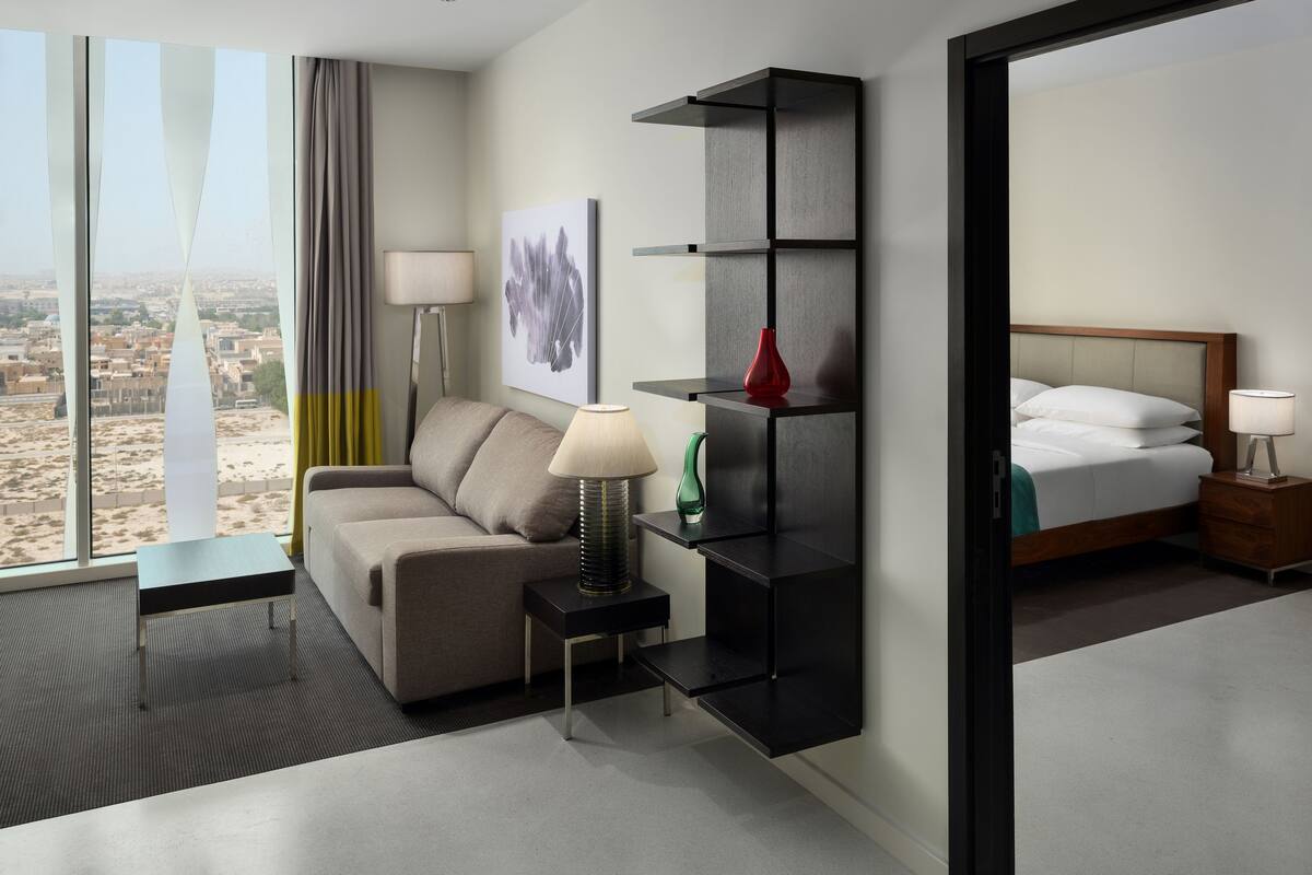 A modern hotel room with a sofa, a small side table with a lamp, a black shelving unit, and a bed with a nightstand.