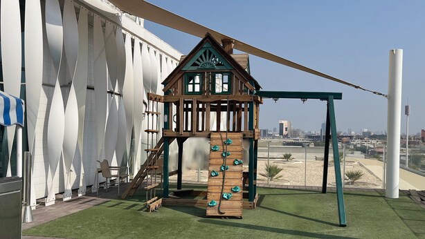 Uma estrutura de recreação infantil com elementos de escalada e baloiço, situada num terraço com vista para a paisagem urbana.