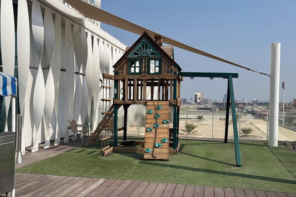 Een speelstructuur voor kinderen met klimelementen en een schommelset, gelegen op een dak met uitzicht op de stad.