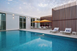 Una piscina con lettini e un tavolo, circondata da un edificio moderno con ampie finestre e un design in legno verticale.