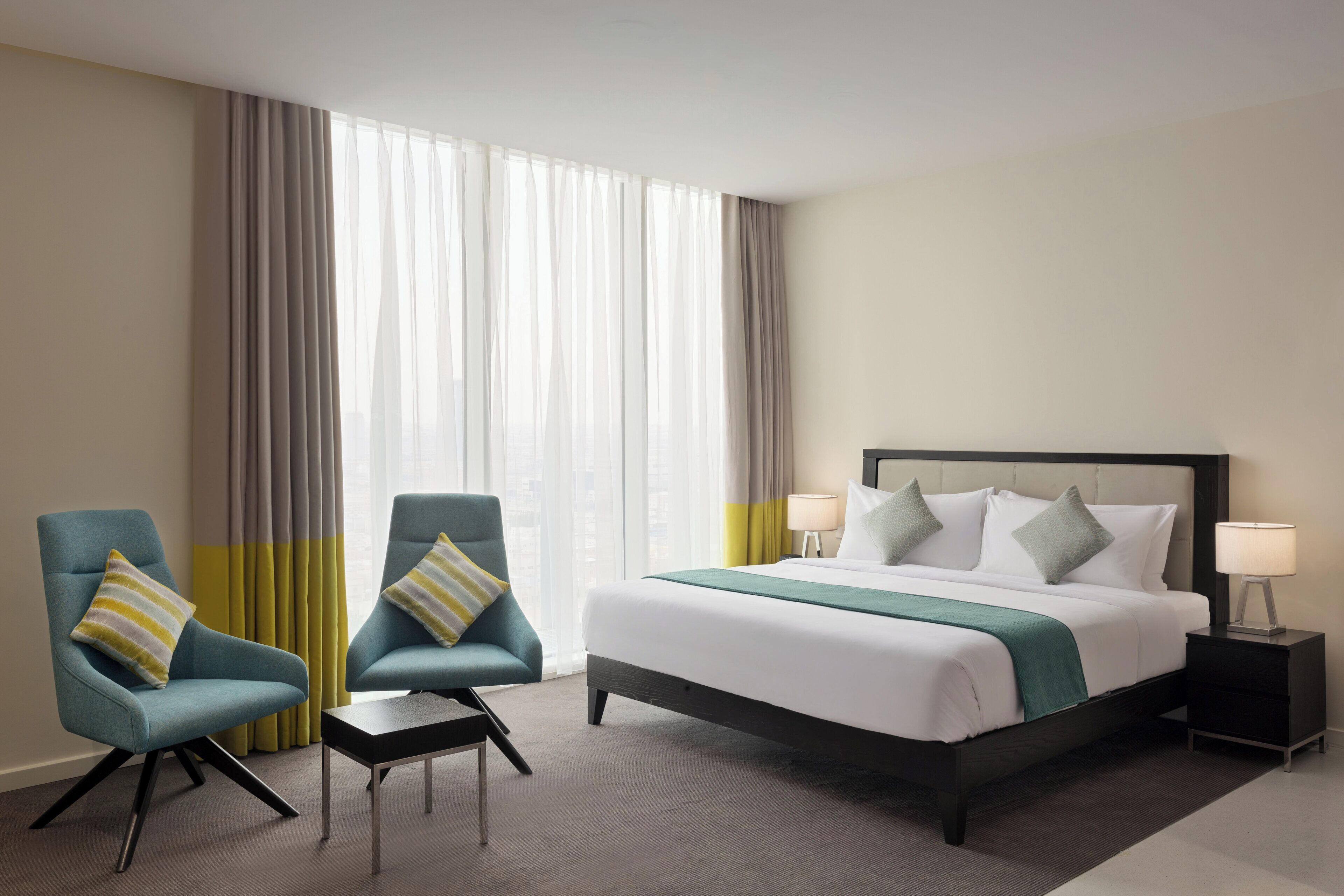 A hotel room with a bed, two armchairs, a small table, a nightstand with a lamp, and sheer curtains.