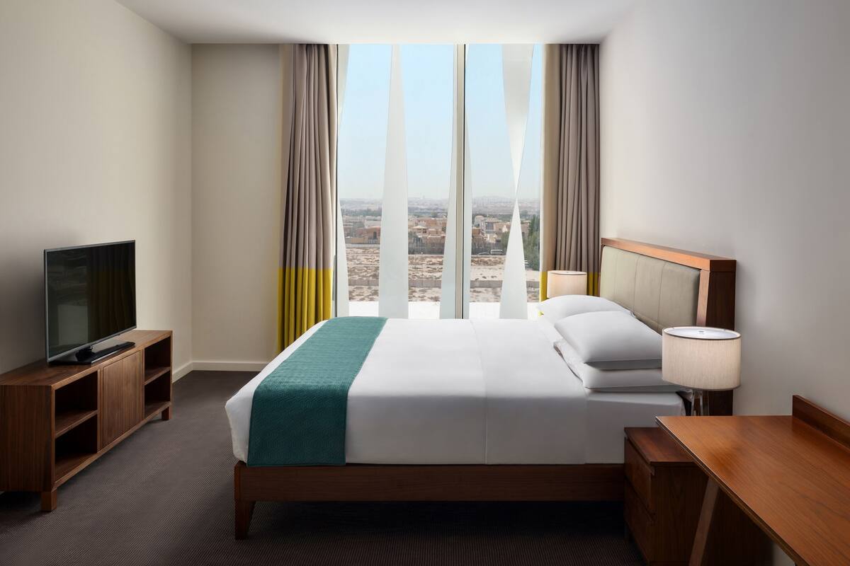 A hotel room with a large bed, a TV on a wooden stand, a desk, and a view of the city through large windows.