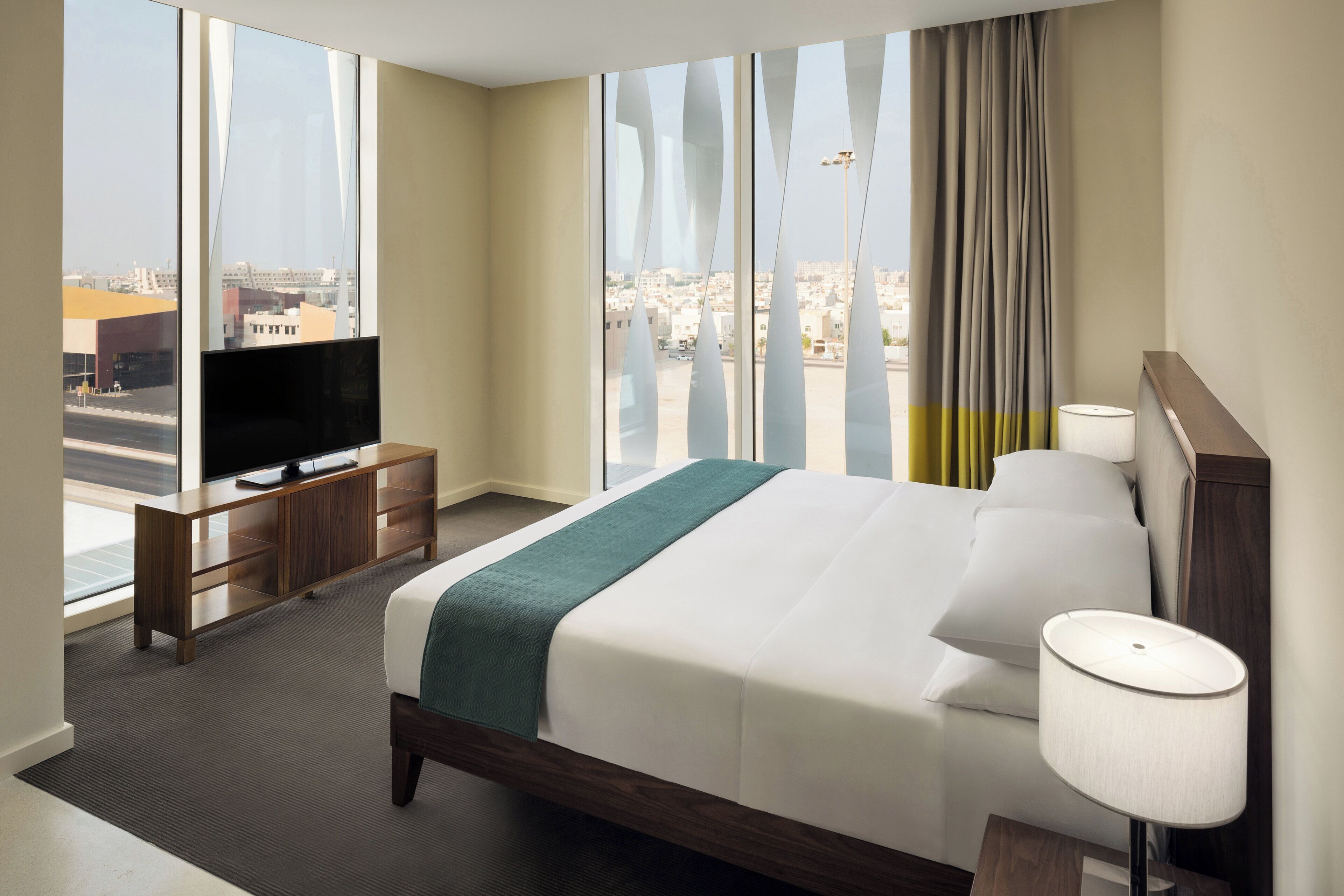 A hotel room with a bed, a TV on a wooden stand, a lamp, and a view of the city through large windows.