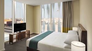 A hotel room with a large bed, a television on a wooden cabinet, and a view of the city through large windows.
