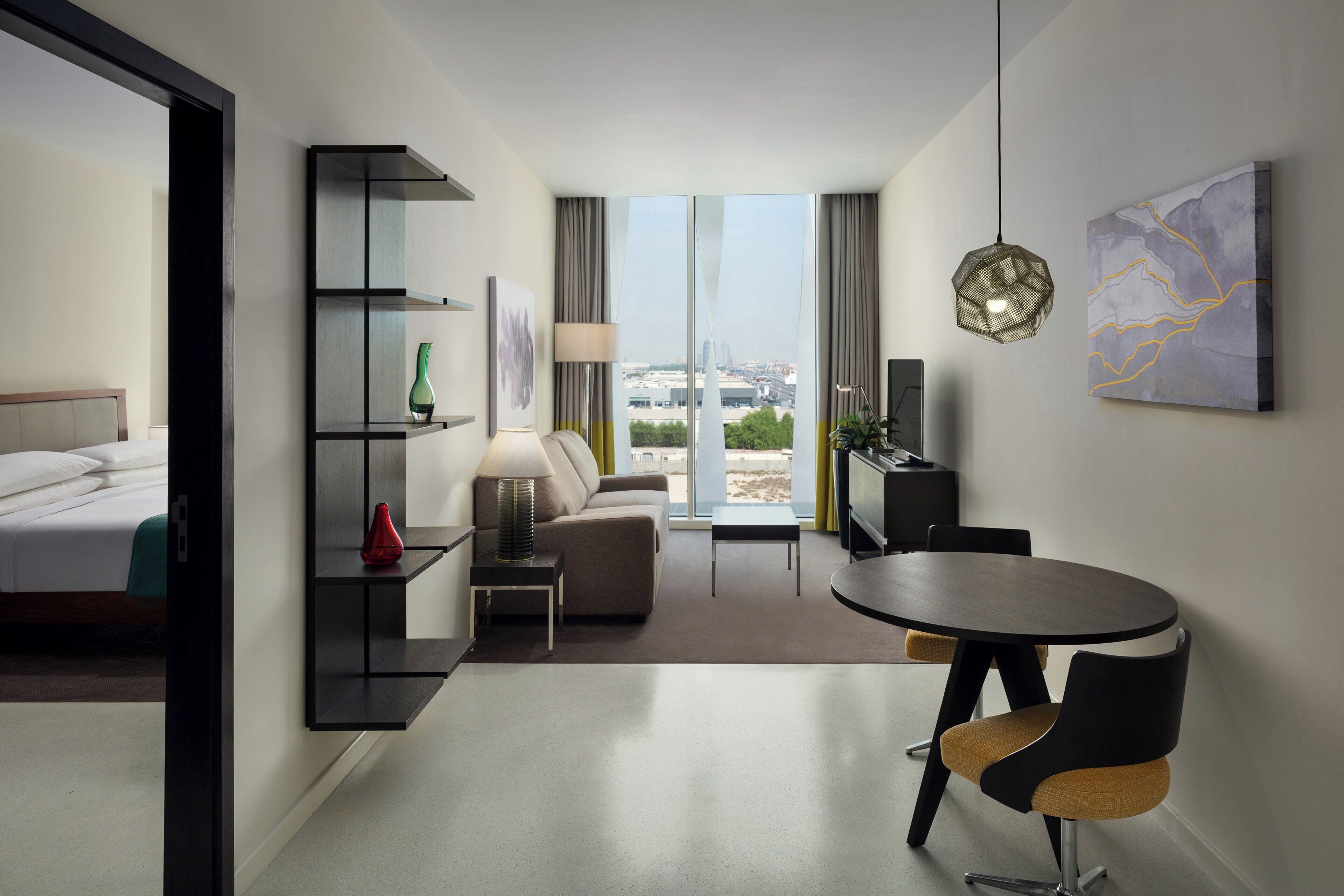A modern hotel room with a bed, a sofa, a dining table with chairs, a TV, and a large window with a city view.