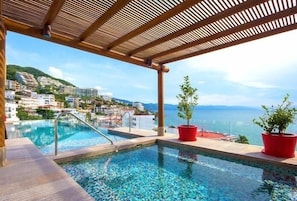 A rooftop infinity pool with a wooden pergola, red planters, and a view of a coastal town.