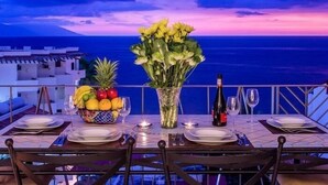 A table set for two with a fruit bowl, a vase of flowers, and a bottle of wine, overlooking a coastal view. - Romantic Zone V177 1 Bedroom (Puerto Vallarta)