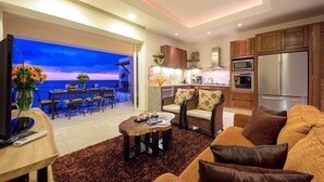 A modern living room with a sofa, coffee table, and TV. There is a dining area with chairs and a table, and a kitchen with cabinets and appliances. - Romantic Zone V177 1 Bedroom (Puerto Vallarta)