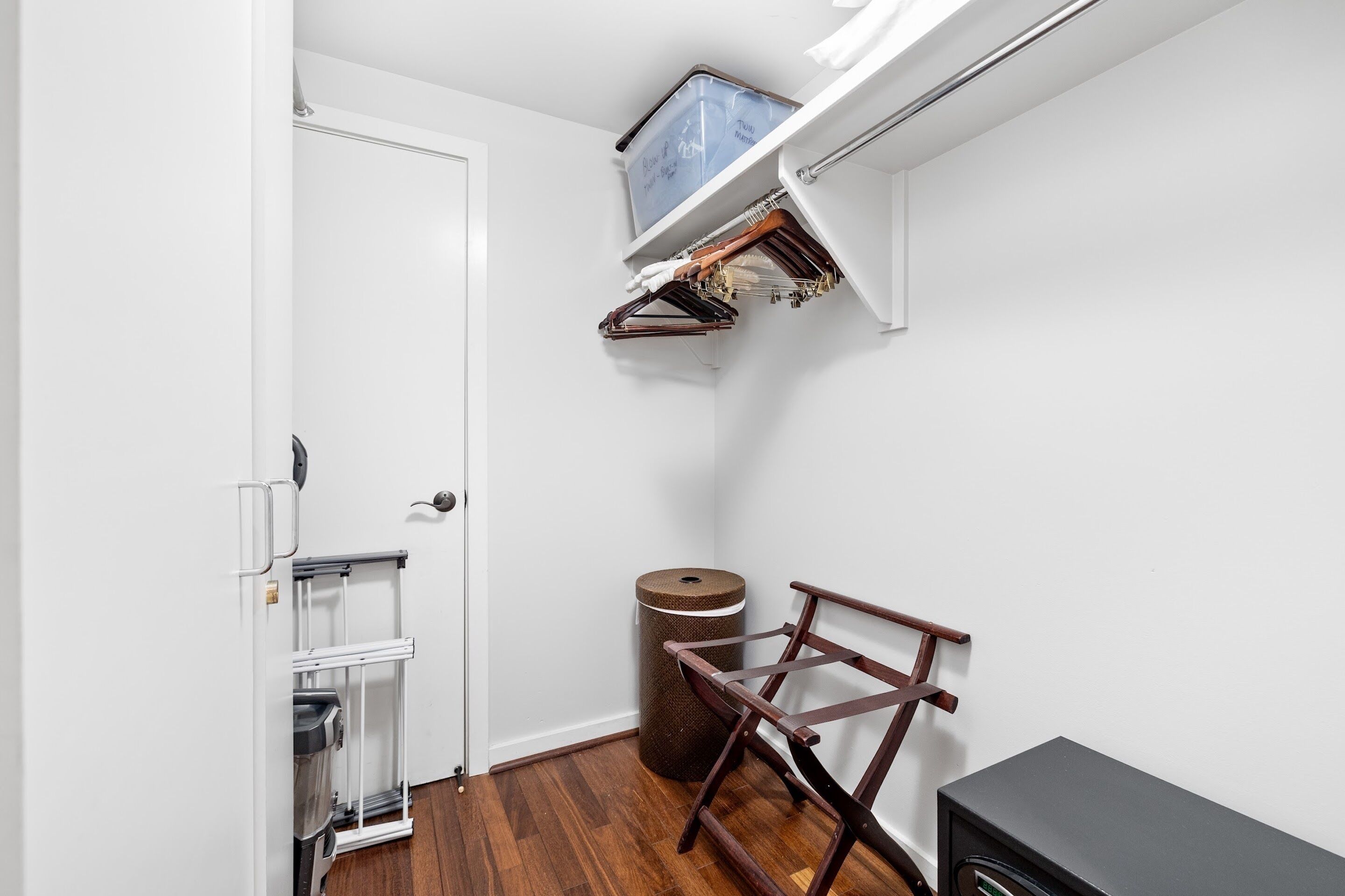 Una habitación pequeña con una puerta blanca, un cesto de basura, un banco de madera y un armario con ropa colgada.
