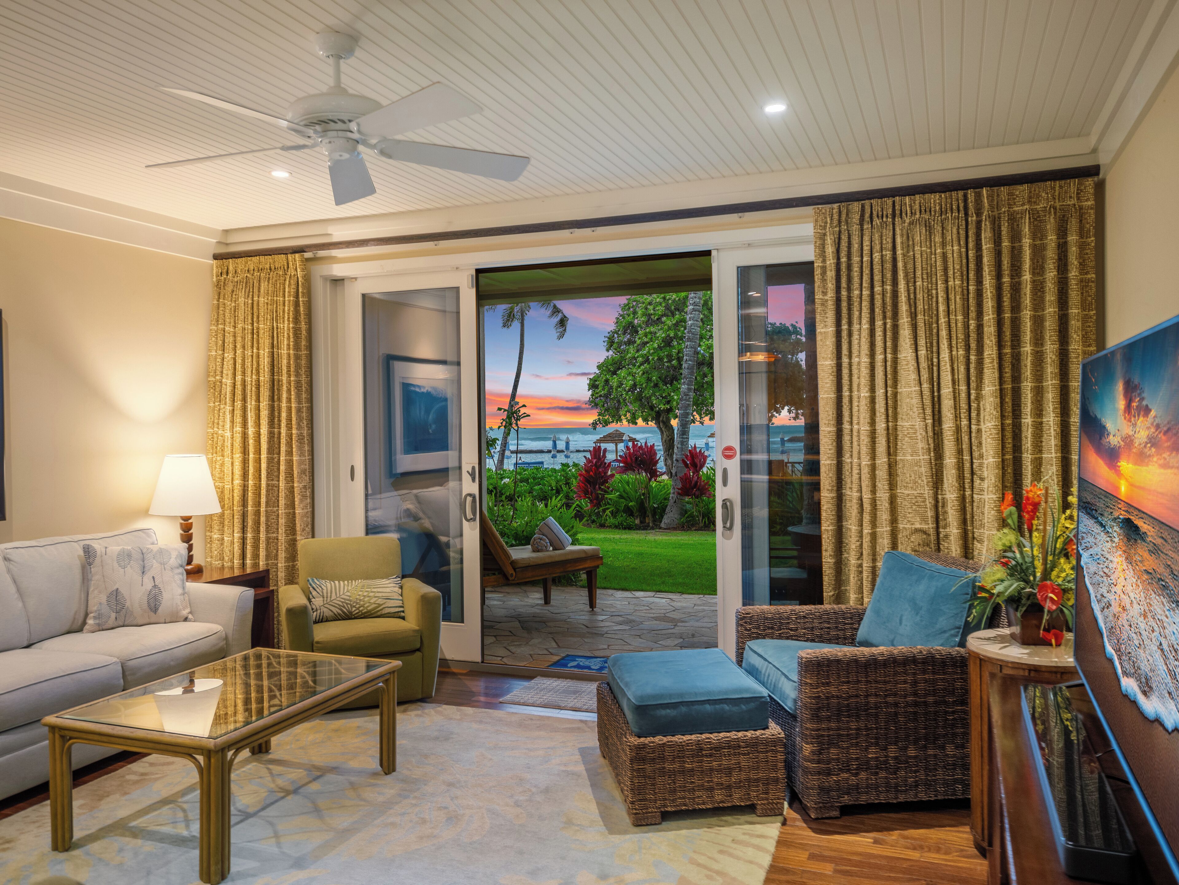 A living room with a glass door leading to a balcony with a view of the ocean and sunset.
