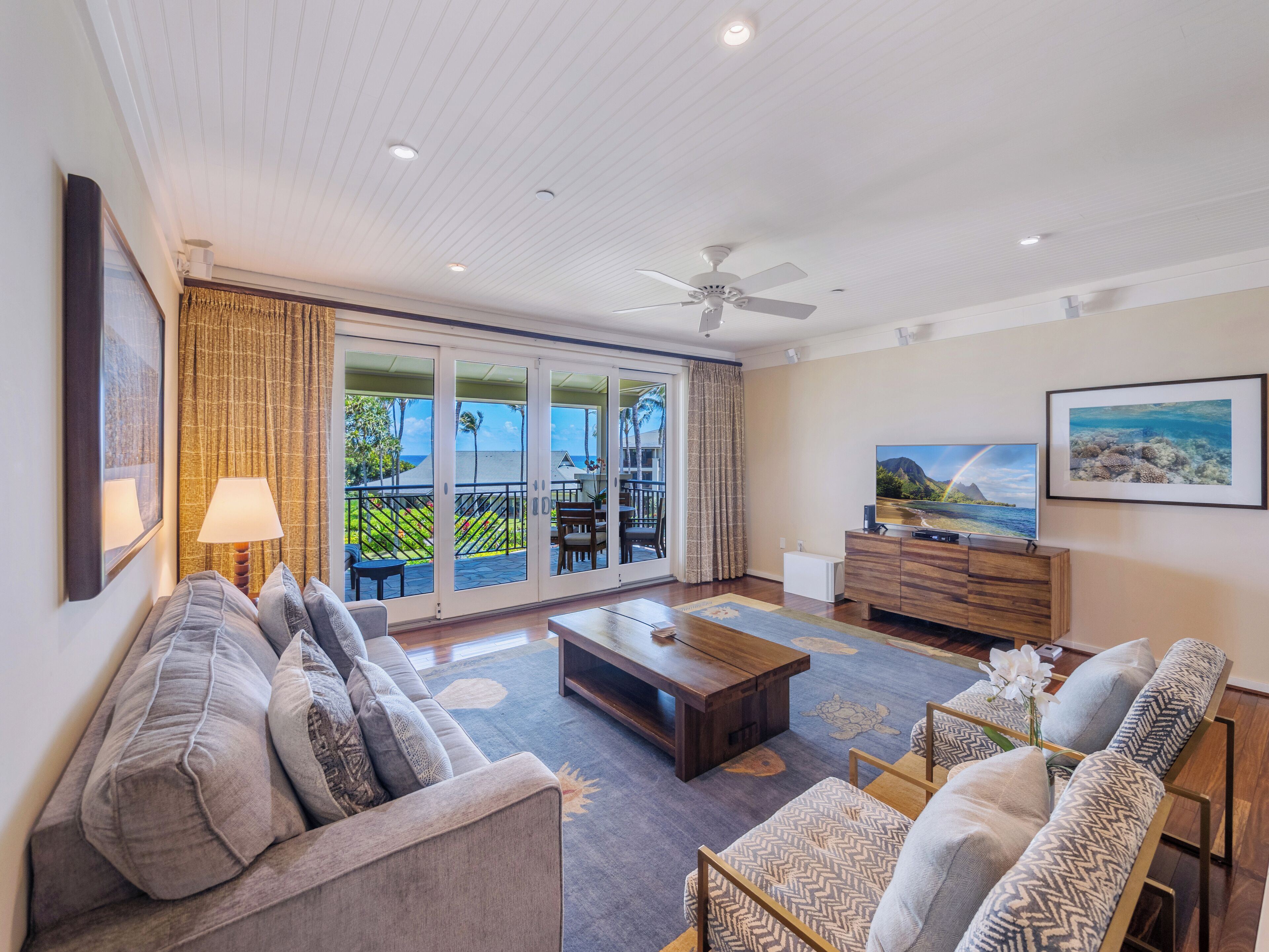 A spacious living room with a sofa, coffee table, and TV, a balcony with sliding doors, and a ceiling fan.