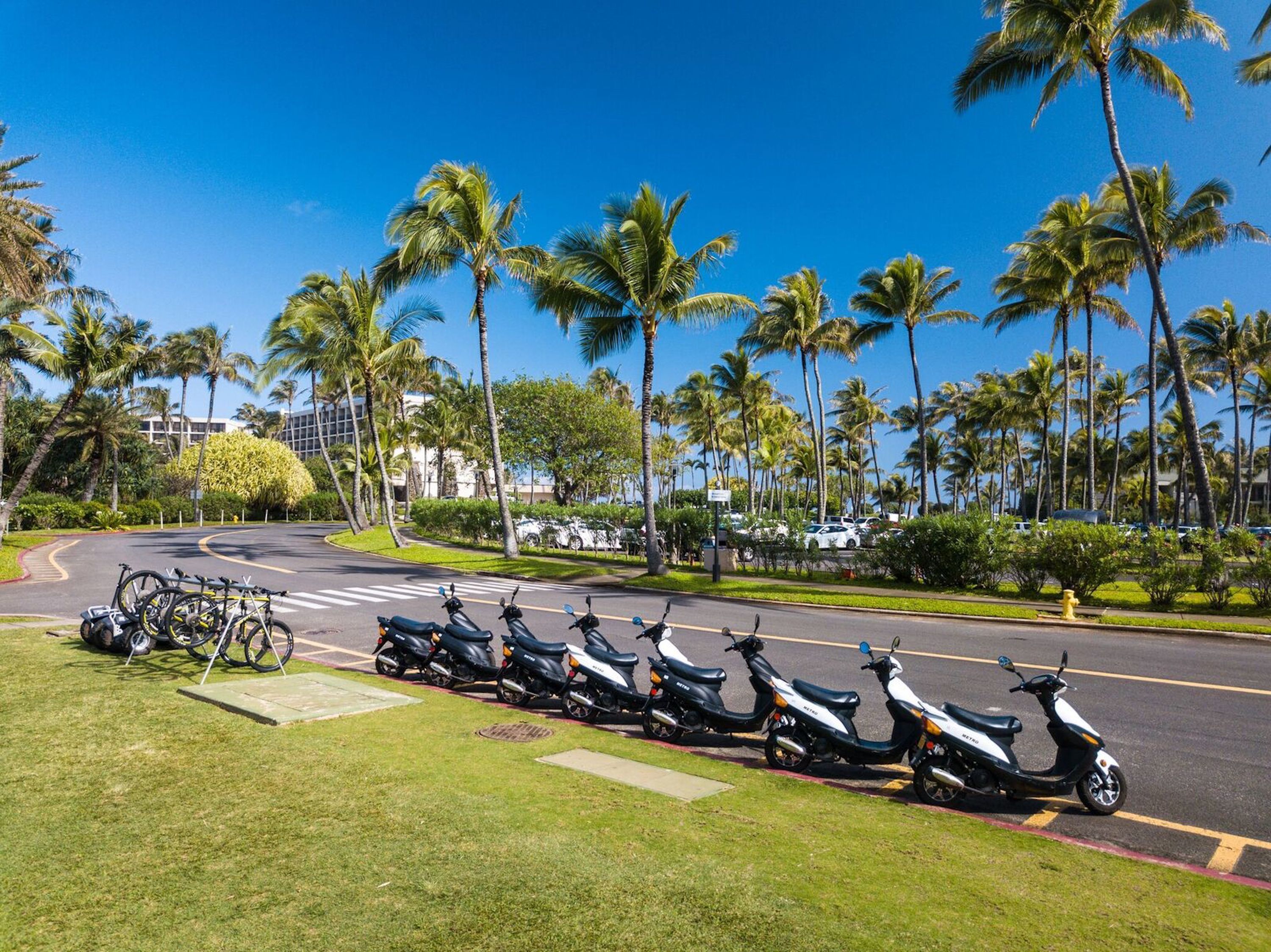 A parking area with several scooters and bicycles, surrounded by palm trees and a clear blue sky.