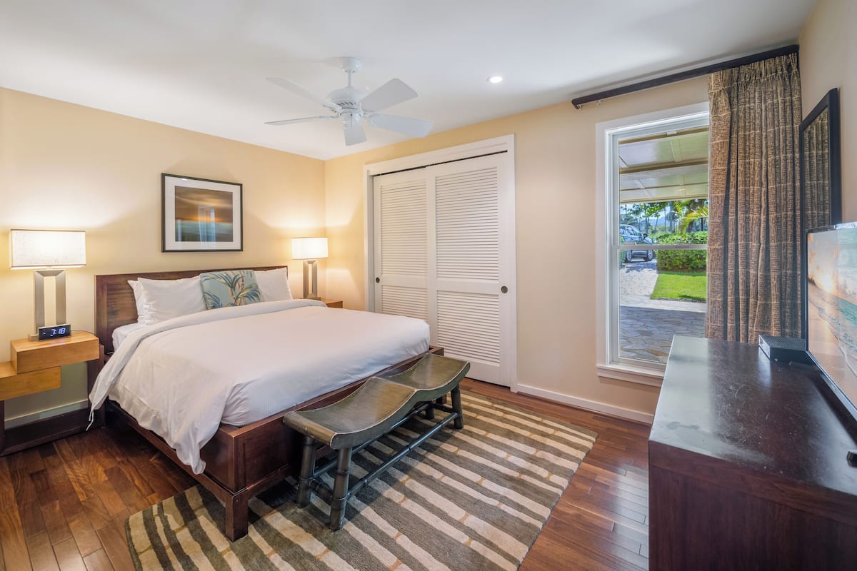 Habitación de hotel con cama, lámparas de noche, ventilador de techo, escritorio y vistas al exterior.