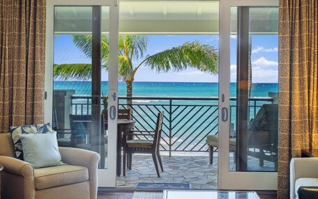 A room with a sliding glass door leading to a balcony with a view of the ocean. There is a chair and a table on the balcony.