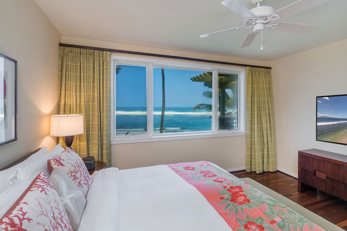 Habitación de hotel con un ventanal que ofrece vistas a la playa y al océano, una cama con ropa de cama floral, una mesita de noche con lámpara, y un televisor sobre un mueble de madera.