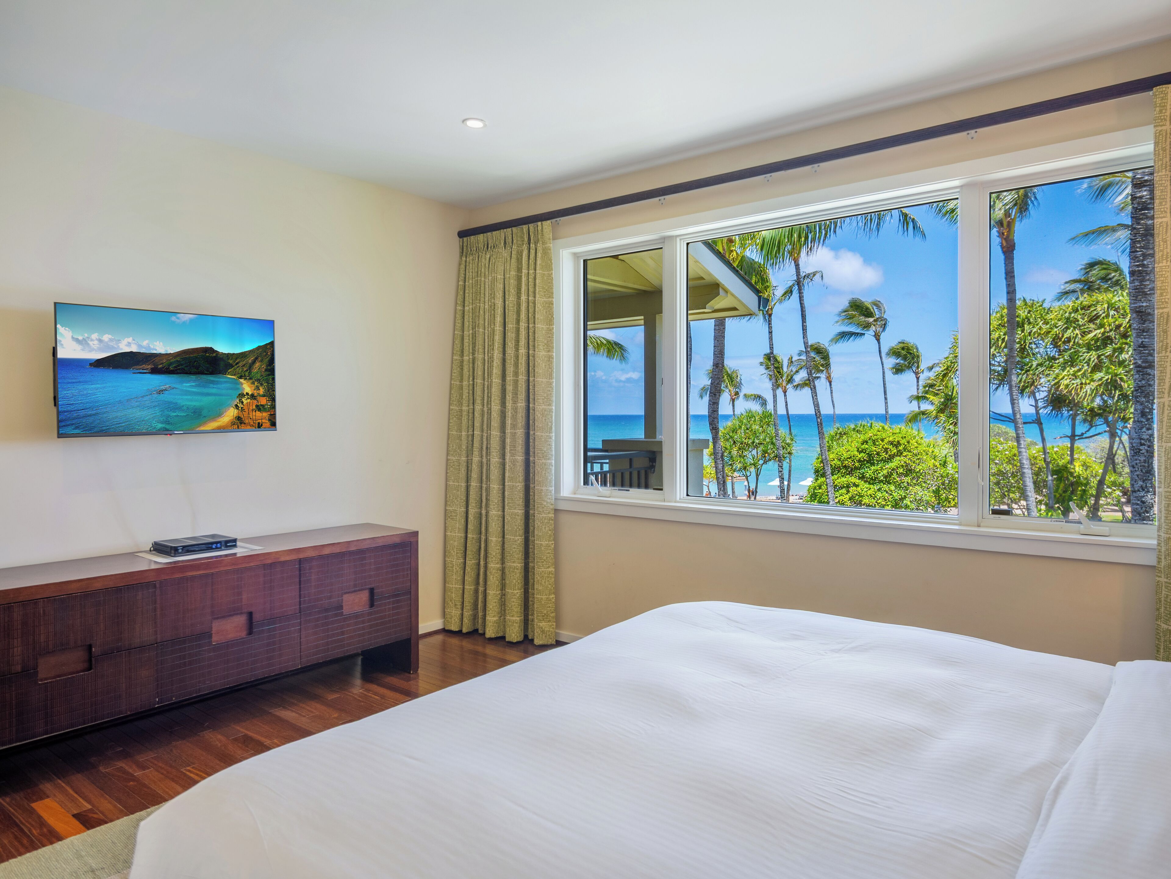 A hotel room with a bed, a TV on the wall, a wooden dresser, and a window with a view of palm trees and the ocean.