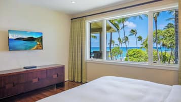 A hotel room with a bed, a TV on the wall, a wooden dresser, and a window with a view of palm trees and the ocean.