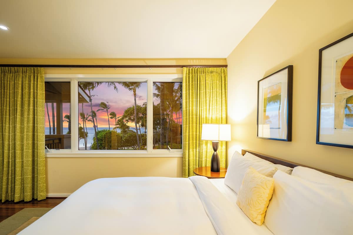 Una habitación de hotel con una cama grande, una mesita de noche con lámpara, una ventana con cortinas y un cuadro en la pared.
