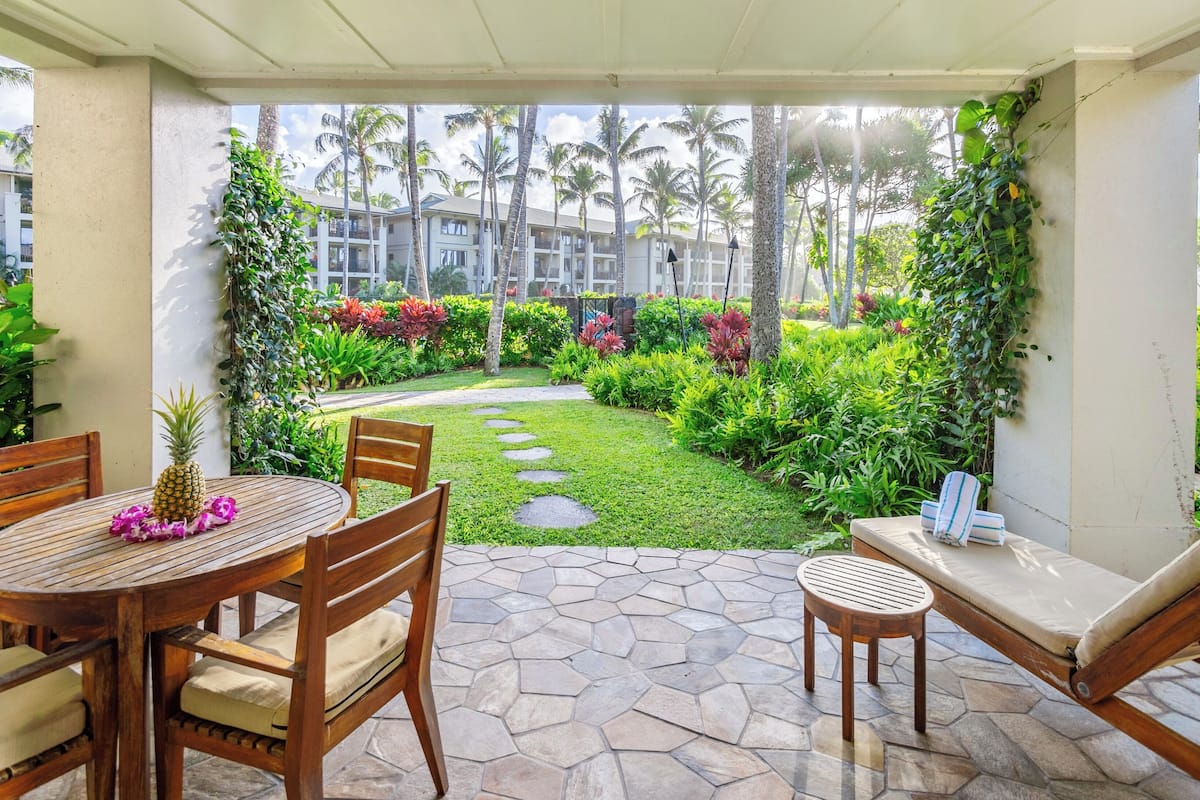 Un patio techado con piso de piedra, muebles de madera y vista a un jardín exuberante con palmeras y plantas tropicales.