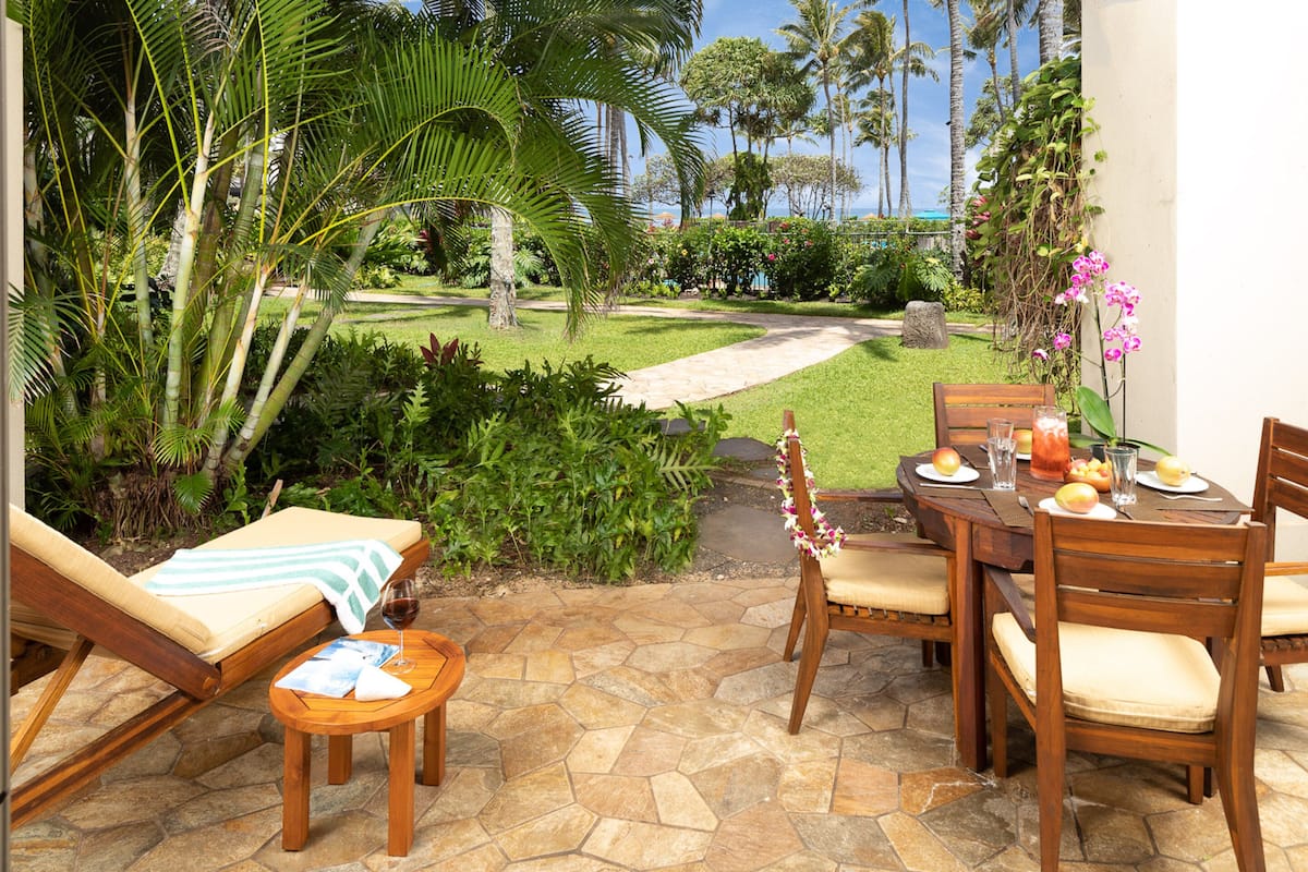 Un patio con piso de piedra, una mesa de madera lista para comer, un sillón de descanso y vistas a un jardín exuberante y palmeras.