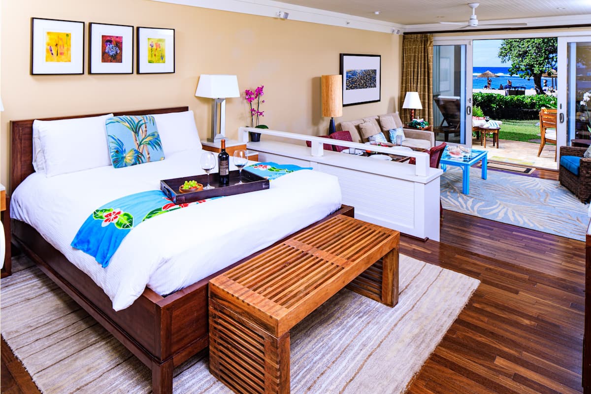 Habitación de hotel con una cama grande, un banco de madera, un sofá y un comedor. Hay cuadros en la pared y vistas a la playa a través de la puerta corredera de vidrio.