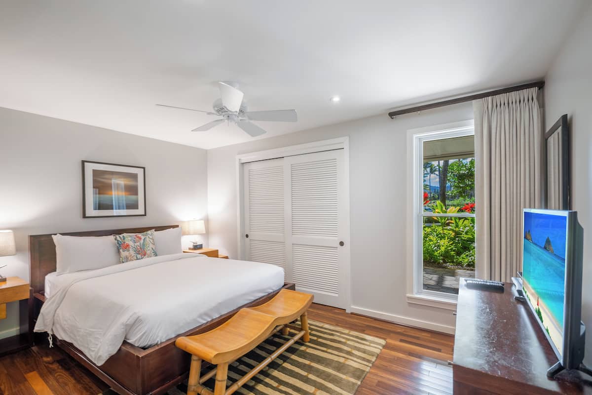 Un dormitorio con una cama, un banco de madera, un ventilador de techo, una mesita de noche, una lámpara, un cuadro enmarcado, una puerta corrediza, una ventana con cortinas y un televisor.