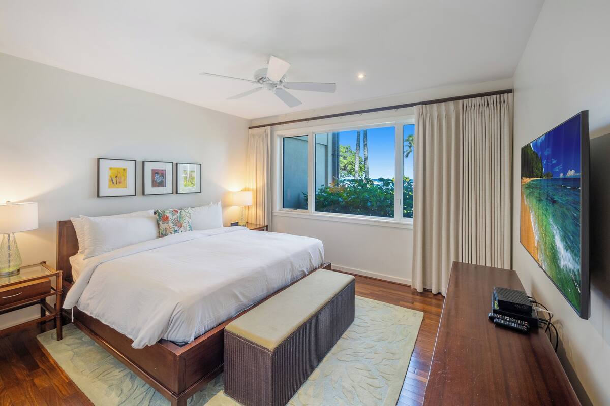 Un dormitorio con cama, ventilador de techo, ventana con cortinas, televisor y cuadros en la pared.