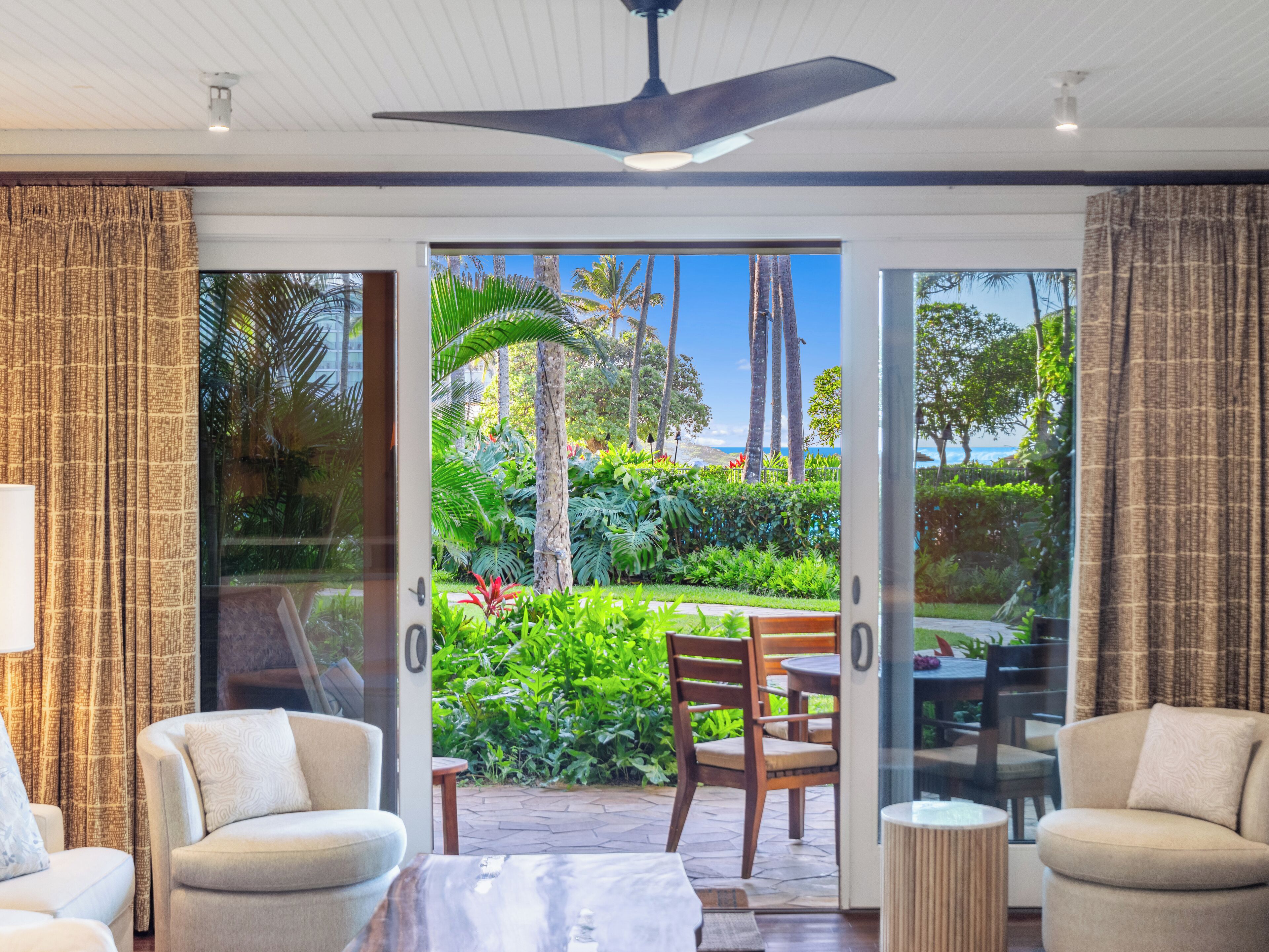 A modern room with a ceiling fan, a glass door leading to a garden with palm trees and a view of the sea.