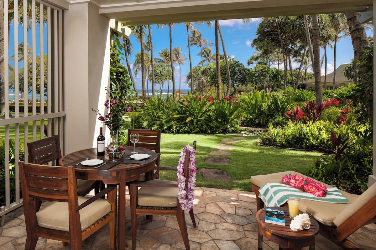 Un patio con una mesa de comedor, sillas y una silla de descanso. Se aprecia una vista de un jardín con plantas tropicales y una vista distante del océano.