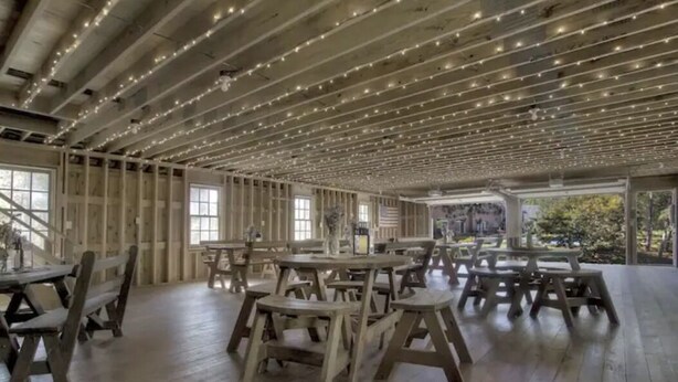 A spacious indoor area with wooden tables and chairs, a high ceiling with string lights, and large windows allowing natural light.