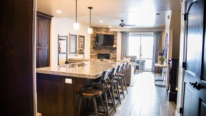 A modern kitchen with a central island, bar stools, a stone fireplace, and a large TV mounted on the wall.