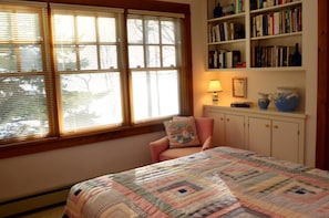 A bedroom with a large window, a chair, a bookshelf, and a quilted bedspread.