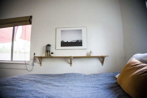 A bedroom with a bed, a window with a view, a shelf with a picture, and a bedside table with a lamp and a plant.