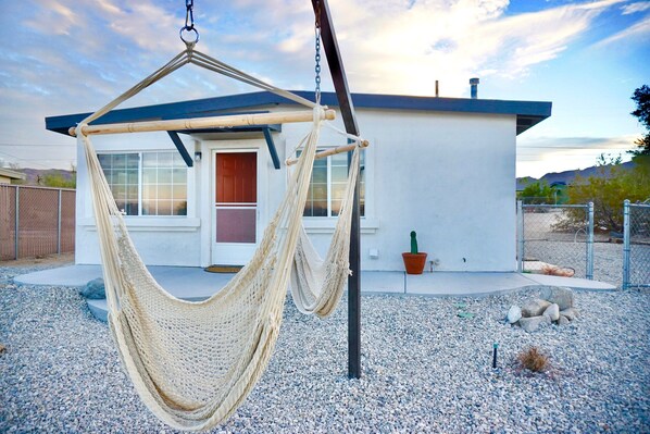 A hammock hanging in front of a house with a gravel driveway and a cactus in the background.