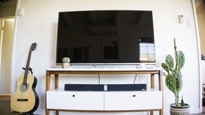 Un televisor de pantalla plana sobre un mueble blanco, con una guitarra y una planta en maceta a un lado.