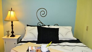 Un lit bien fait, recouvert d’une couvre-lit à motifs noir et blanc, avec un oreiller noir et un oreiller blanc orné de motifs de feuilles. Une petite table de chevet, sur laquelle se trouvent une lampe et un objet décoratif, est placée à côté du lit. Un magazine et une tasse avec une paille sont posés sur le lit.